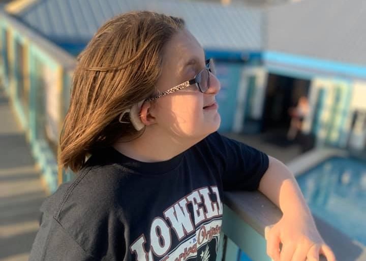 A 13-year-old girl with deaf-blindness outside with her hand on a railing. A peaceful look on her face. She has a cochlear implant and is wearing glasses.